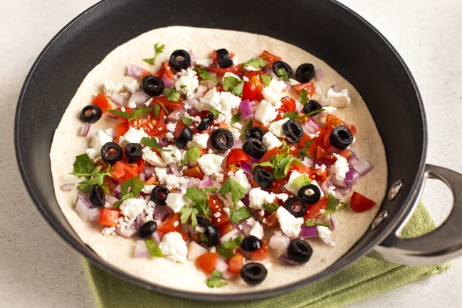 Greek quesadilla before cooking - a tortilla in a pan topped with feta, olives, tomatoes, red onion and parsley