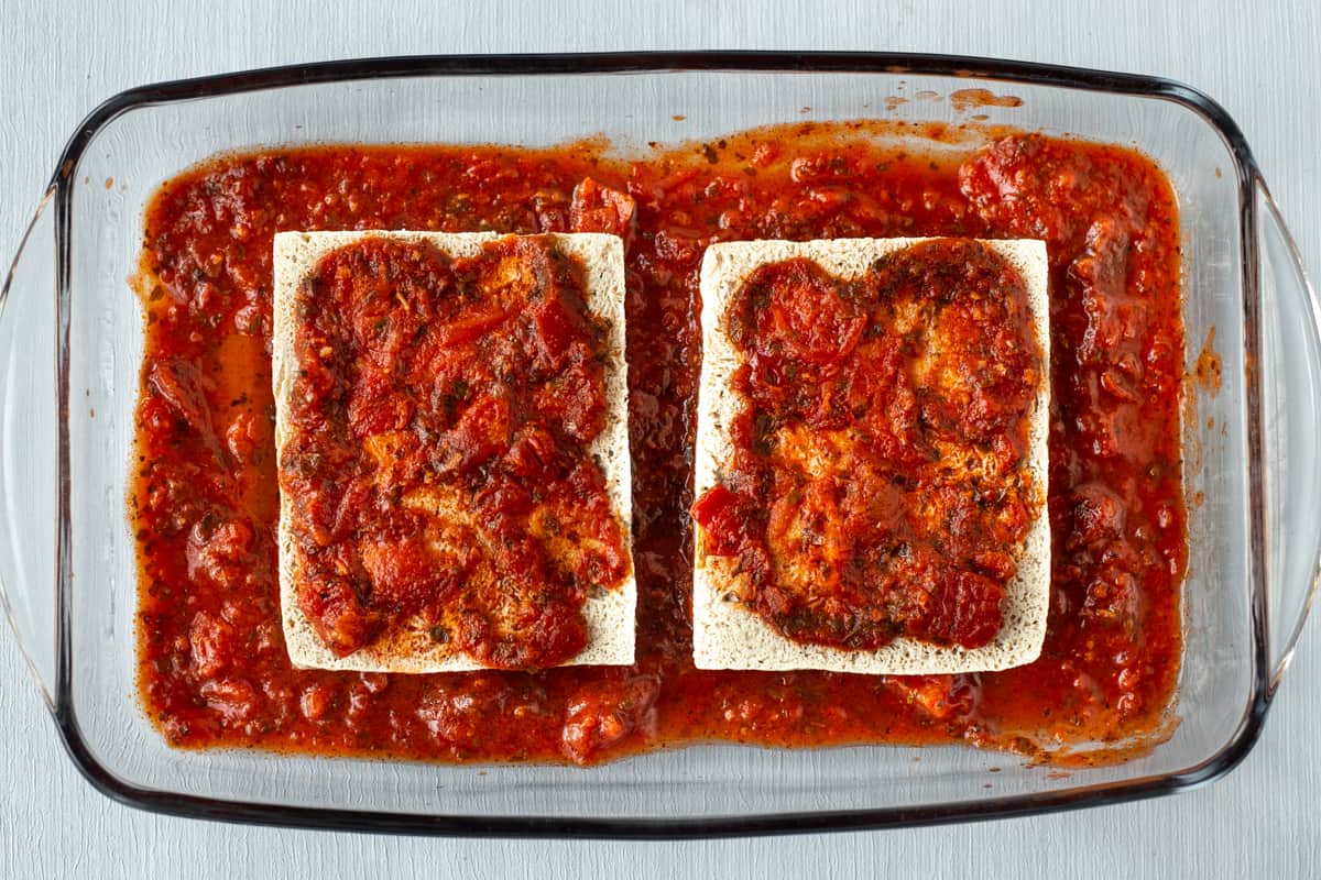 Two slices of tofu in a baking dish with tomato sauce.