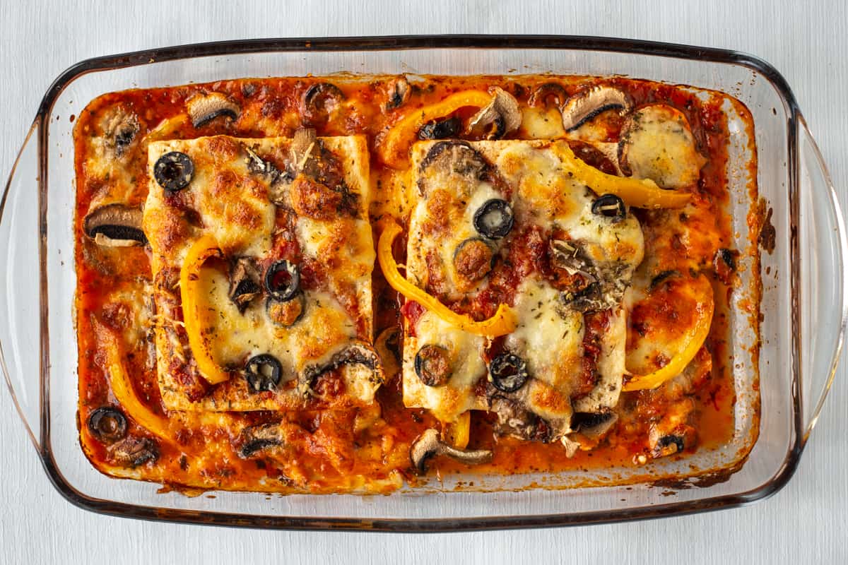 Tofu in a baking dish, with tomato sauce, pizza toppings, and crispy cheese.