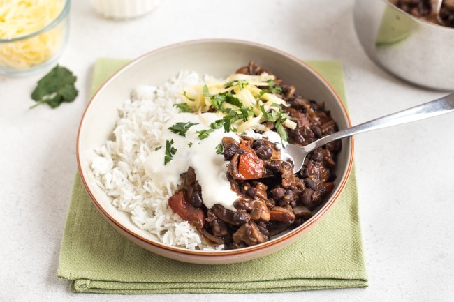 Mushroom and black bean chilli in a bowl with rice and sour cream, with a scoop being taken with a fork