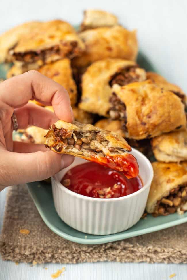 A hand holding up a vegetarian sausage roll dipped in ketchup, with the lentil filling showing.