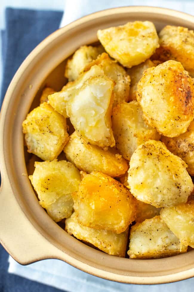 A close up of a dish full of roast potatoes, with one broken in half, showing the fluffy centre.