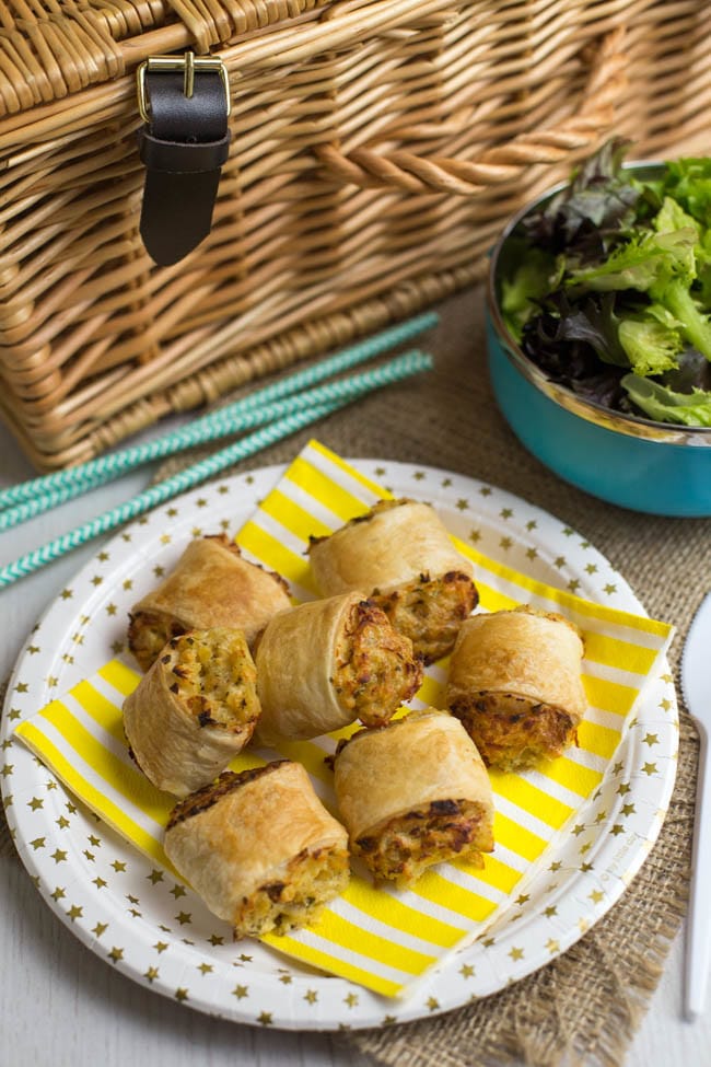 A few vegetarian sausage rolls on a paper plate with a picnic basket.