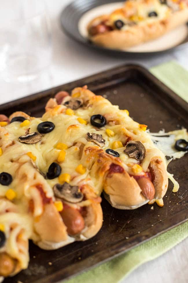 Pizza hot dogs in a row topped with mushrooms, sweetcorn and black olives.