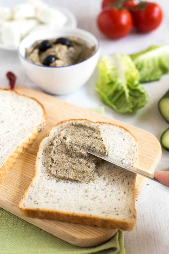 Homemade black olive hummus being spread on a piece of bread.