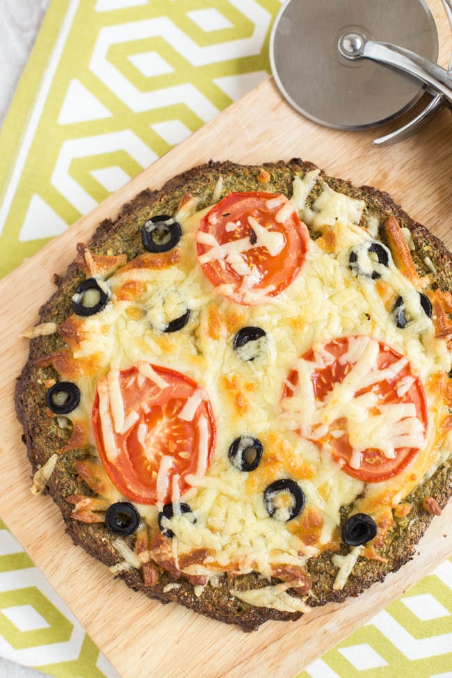 A cheesy broccoli crust pizza on a wooden board, topped with sliced tomatoes and olives.