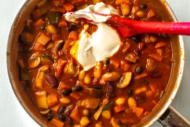 A dollop of sour cream being added to a rich tomatoey bean stew in a saucepan.