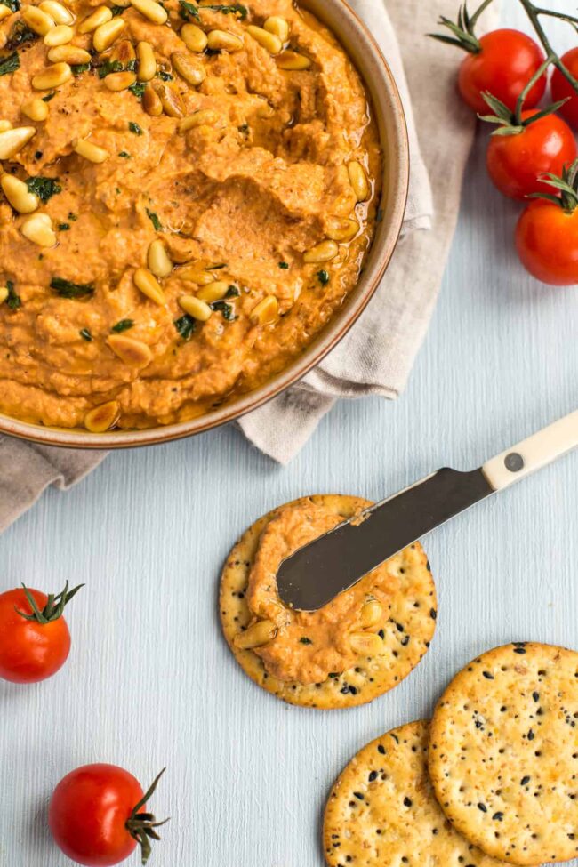 A knife spreading tomato hummus on a cracker.