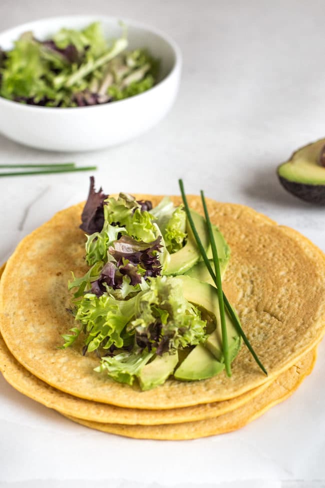 A stack of gluten-free lentil tortillas topped with lettuce and avocado, with a bowl of lettuce in the background