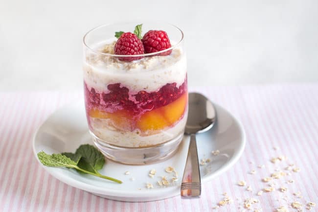 Layered overnight oats in a glass on a saucer, with peaches and raspberry sauce, topped with fresh raspberries and mint