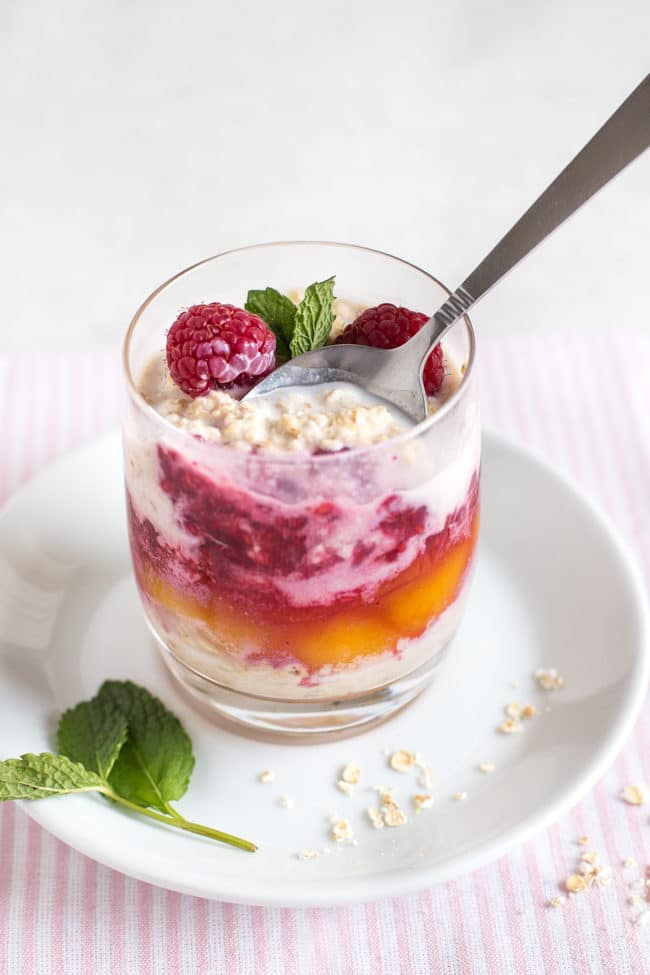 Layered overnight oats with peaches and raspberry sauce, topped with fresh raspberries and mint, with a spoon