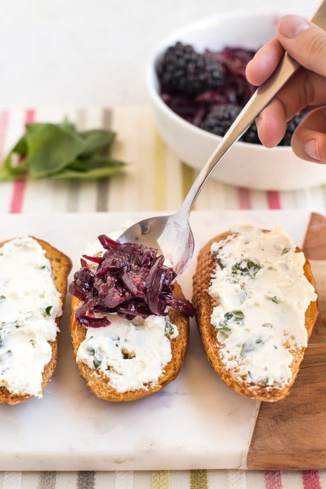 A spoonful of balsamic blackberry compote being spooned onto goat's cheese crostini