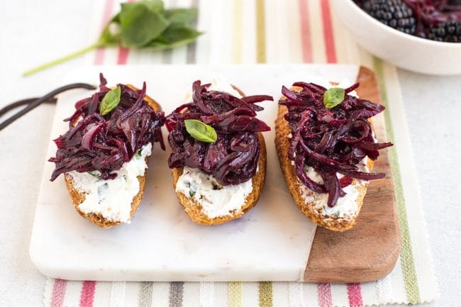 Three pieces of goat's cheese crostini with red onion and blackberry compote, on a white board