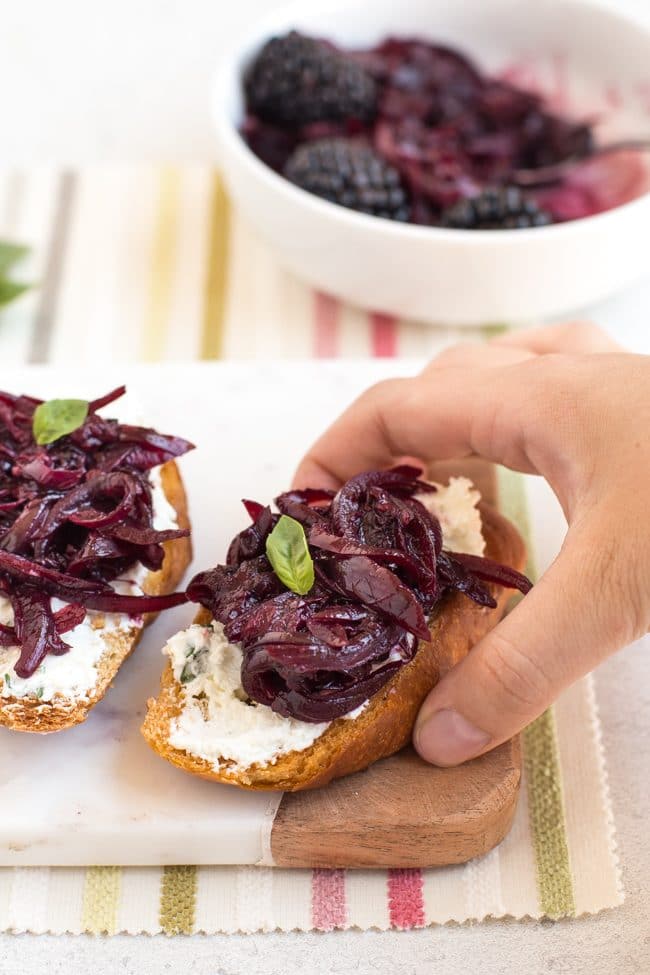 A hand taking a piece of goat's cheese crostini with red onion and blackberry compote