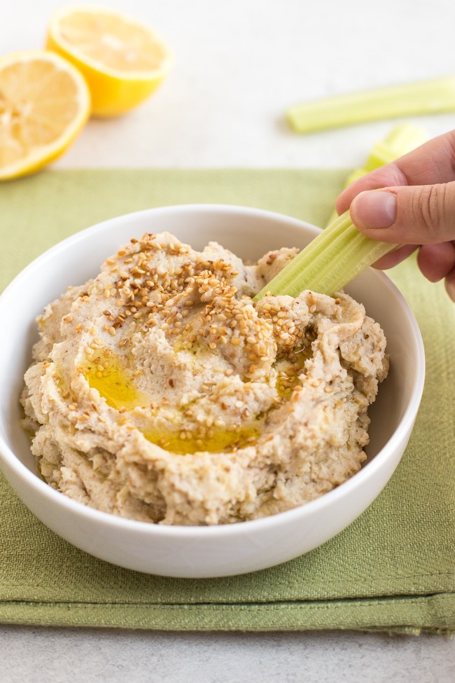 Low-carb cauliflower hummus in a bowl, with a hand taking a scoop with a celery stick