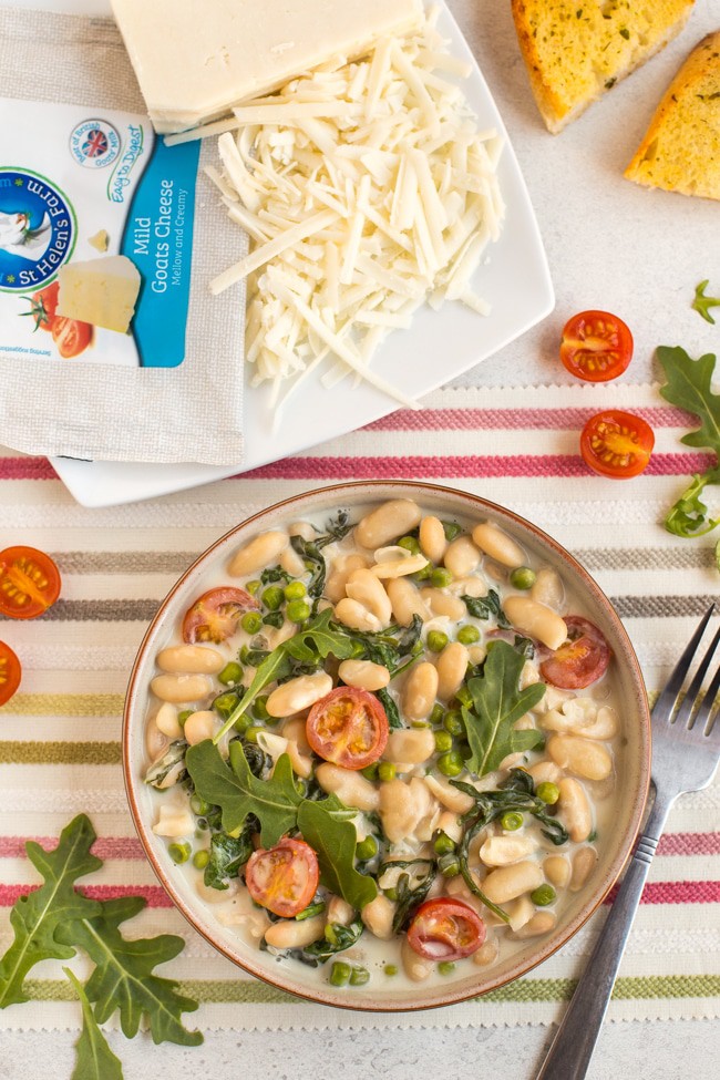 Creamy white beans with rocket and tomatoes in a bowl with a plate of grated goat's cheese