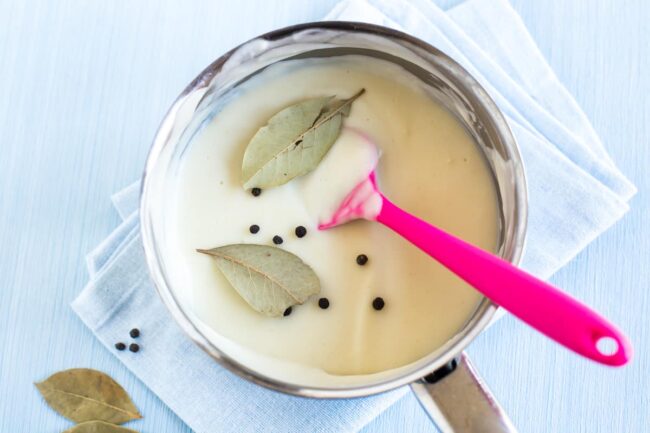 A creamy white sauce in a saucepan with added bay leaves and whole peppercorns