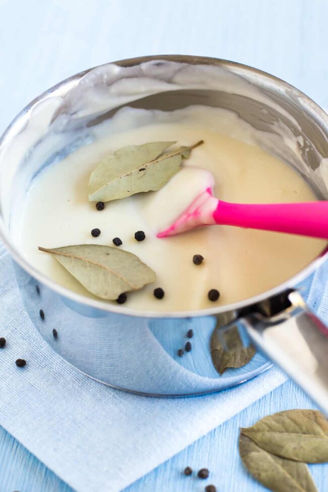 Creamy white sauce in a saucepan with bay leaves and whole peppercorns