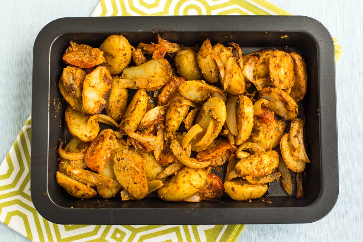 Roasted Bombay potatoes with tomatoes and onions on a baking tray.