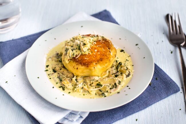 A crispy mashed potato cake on a plate, drizzled with a creamy mushroom sauce and sprinkled with cheese.