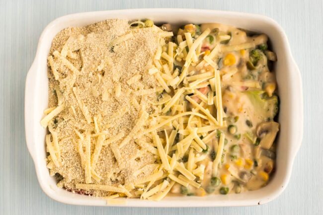 Creamy vegetables in a baking dish, being topped with grated cheese and breadcrumbs.