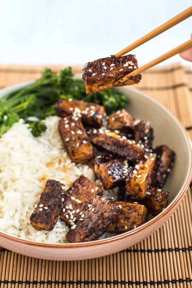 A pair of chopsticks picking up a piece of sticky teriyaki tofu from a bowl.