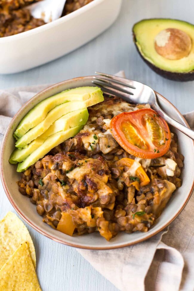 Portion of enchilada lentil casserole in a bowl with sliced tomato and avocado.