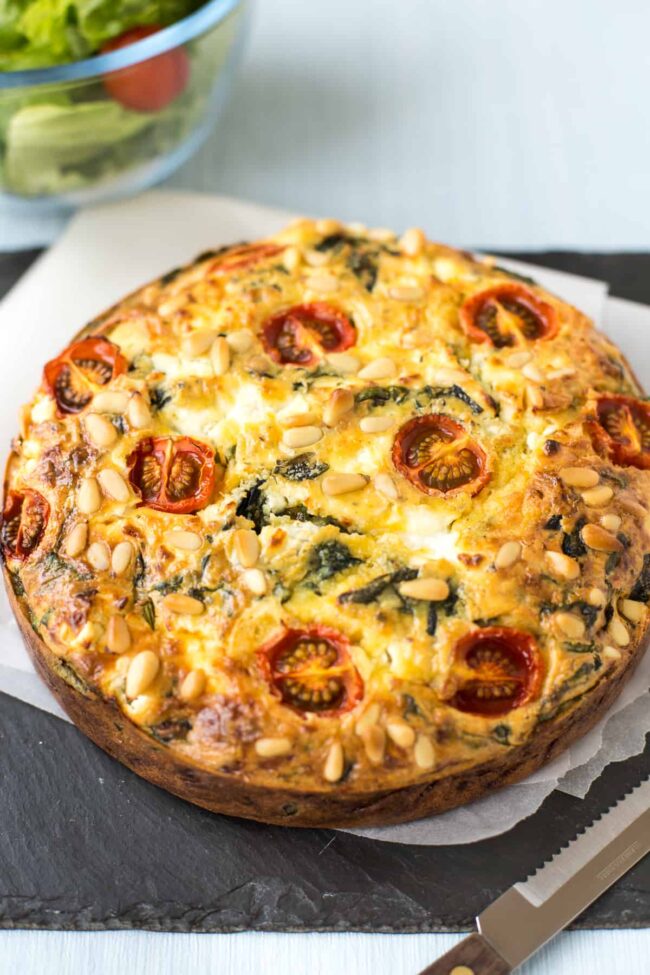 A spinach and goat's cheese quiche on a board, topped with cherry tomatoes and pine nuts.