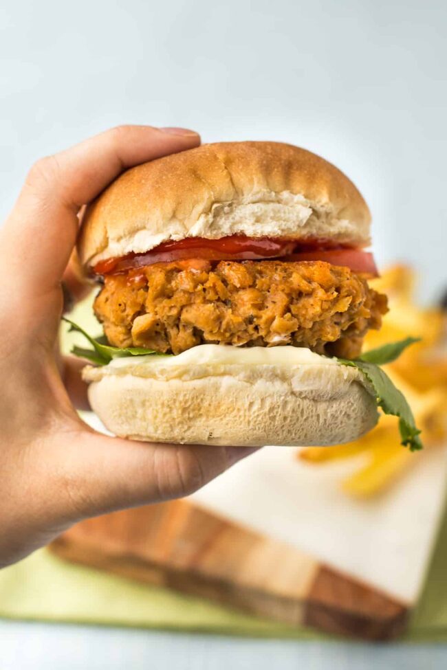 A hand holding up a vegan chicken burger in a bun with lettuce, tomato, ketchup and mayo.