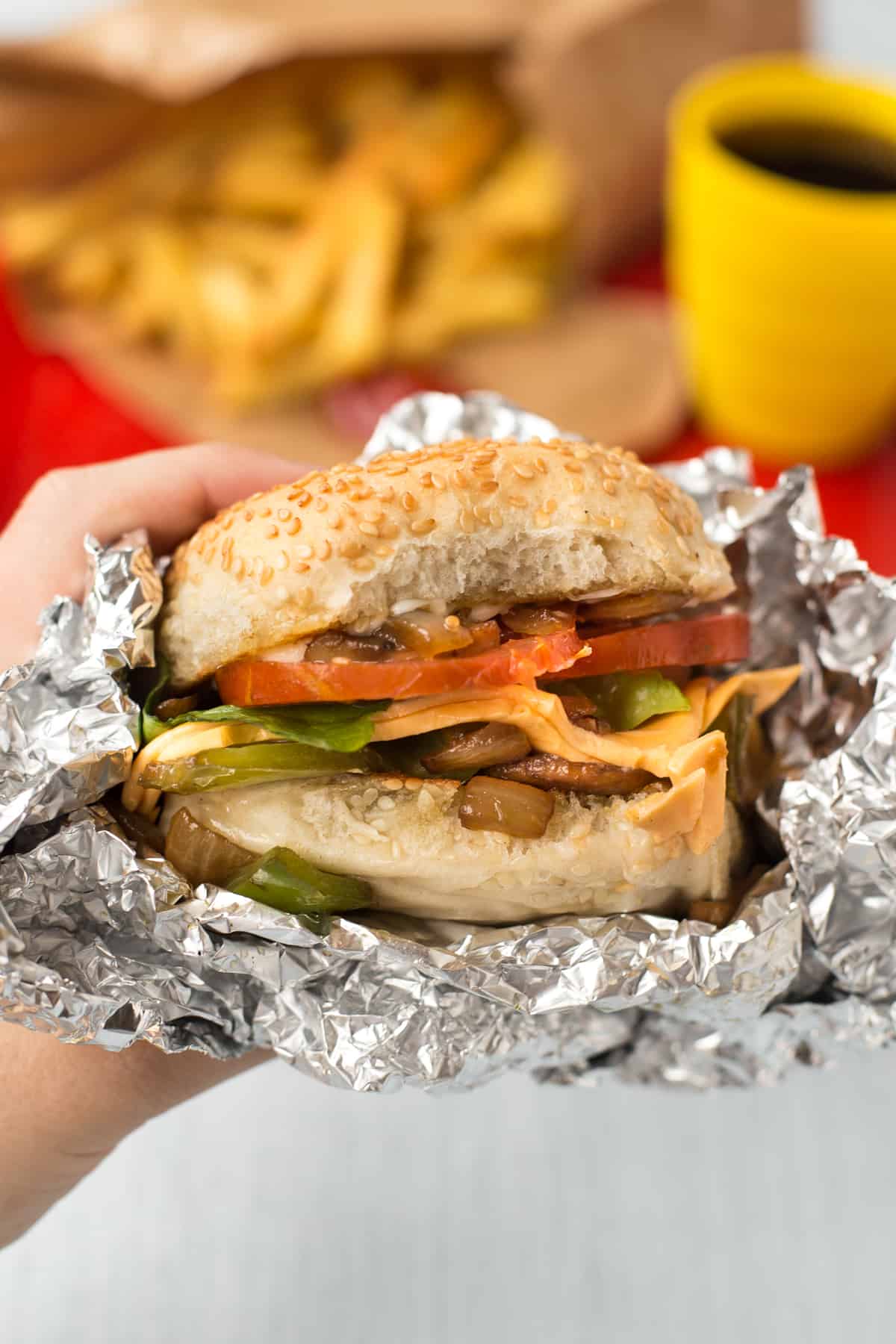 A hand holding up a homemade Five Guys cheese veggie sandwich wrapped in foil.