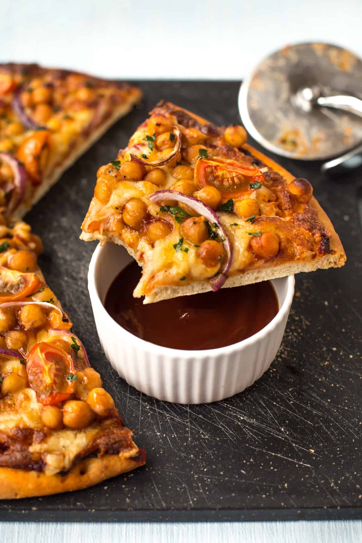 A slice of BBQ chickpea pizza with a bite taken, resting on top of a ramekin of BBQ sauce.