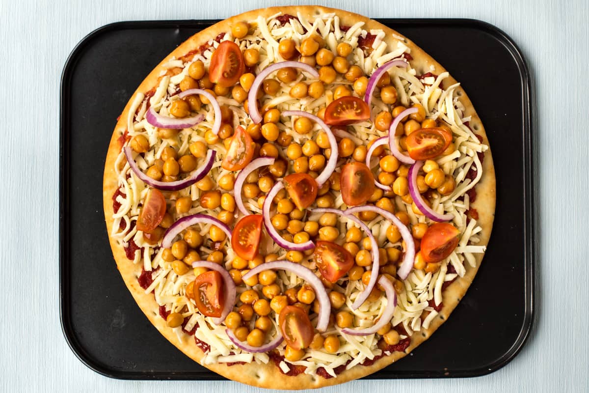 An uncooked pizza topped with BBQ chickpeas, red onion and cherry tomatoes.