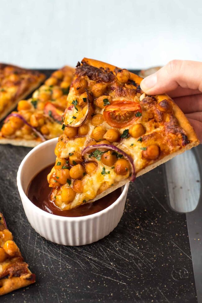 A slice of BBQ chickpea pizza being dipped into a ramekin of BBQ sauce.
