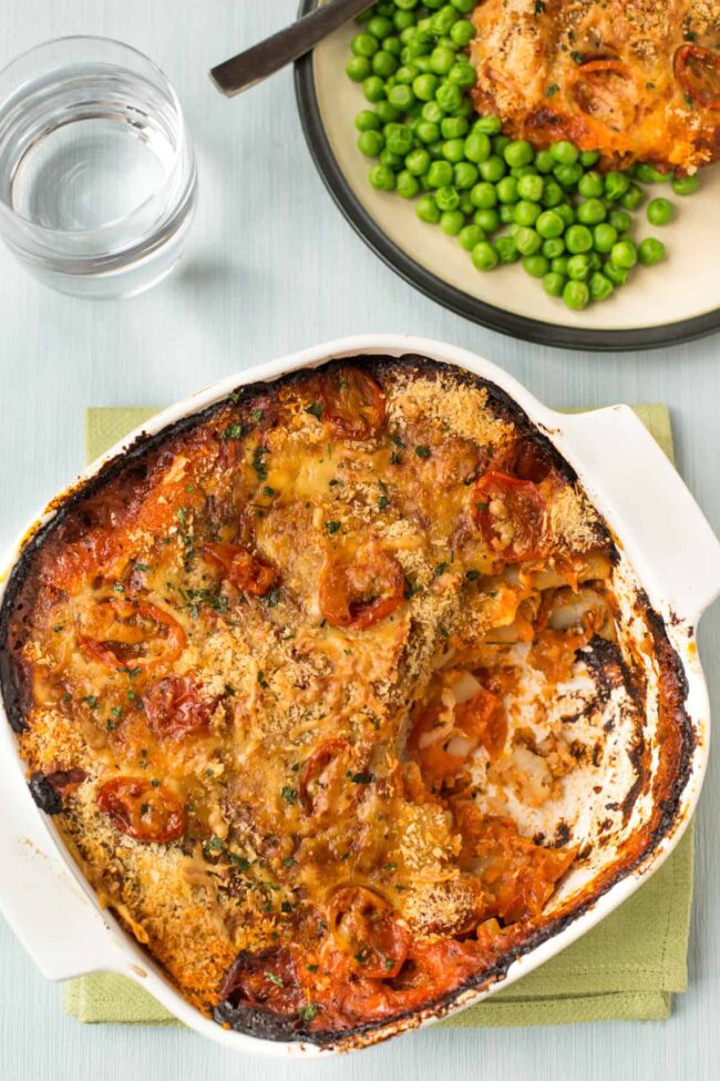 Cheese and tomato potato bake in a dish with a scoop removed, seen behind served with peas.