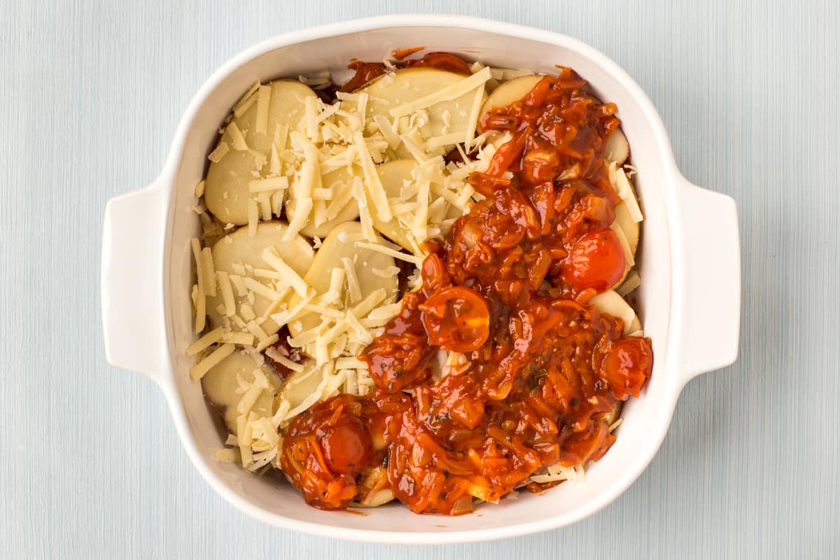 Uncooked potato bake being layered in a dish with tomato sauce and cheese.
