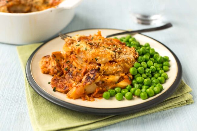 A portion of cheese and tomato potato bake on a plate with green peas.