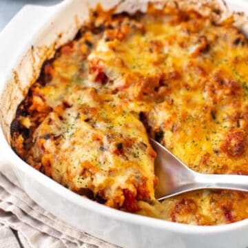 A large spoon taking a scoop from a baking dish full of cheesy tomato baked rice.