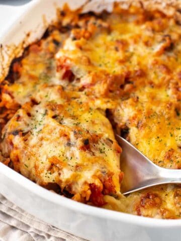 A large spoon taking a scoop from a baking dish full of cheesy tomato baked rice.