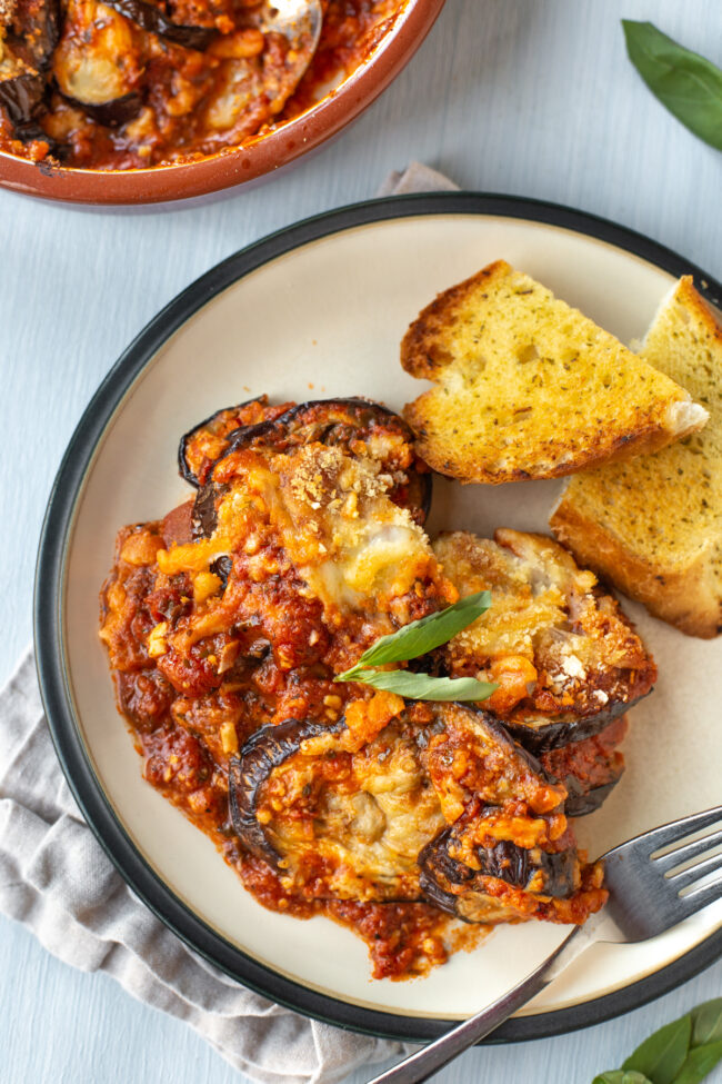 A portion of cheesy eggplant bake on a plate with garlic bread.