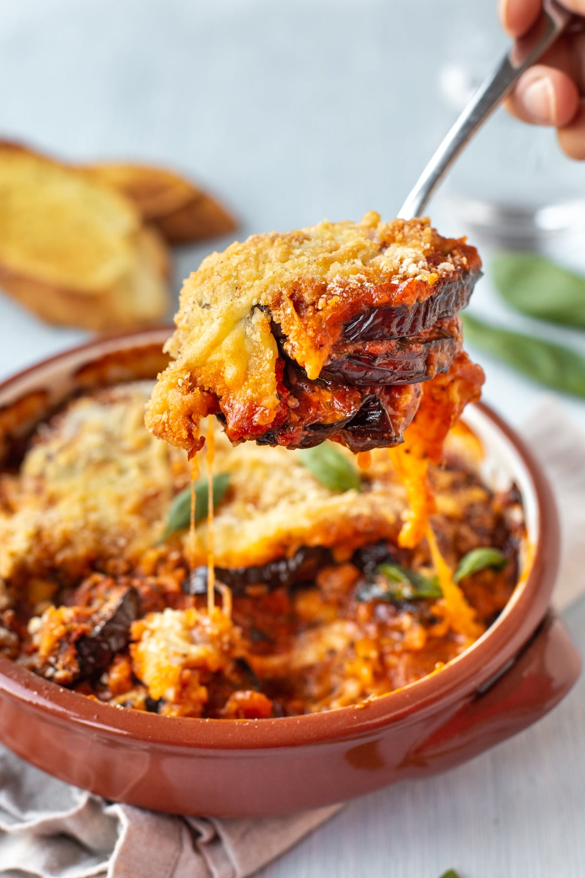 A spoon scooping some cheesy eggplant bake from a dish, with trailing strings of stretchy, gooey mozzarella.