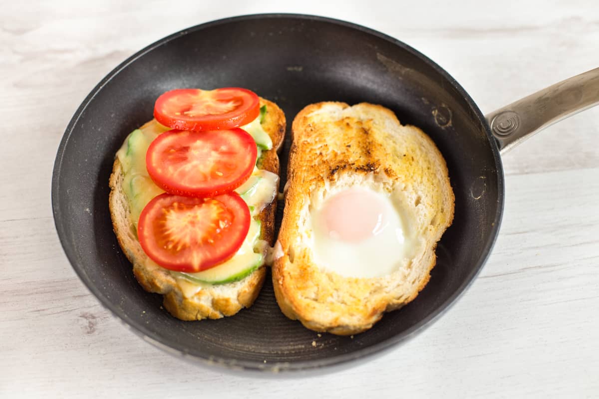 Egg in a hole in a frying pan with another piece of cheesy tomato toast.