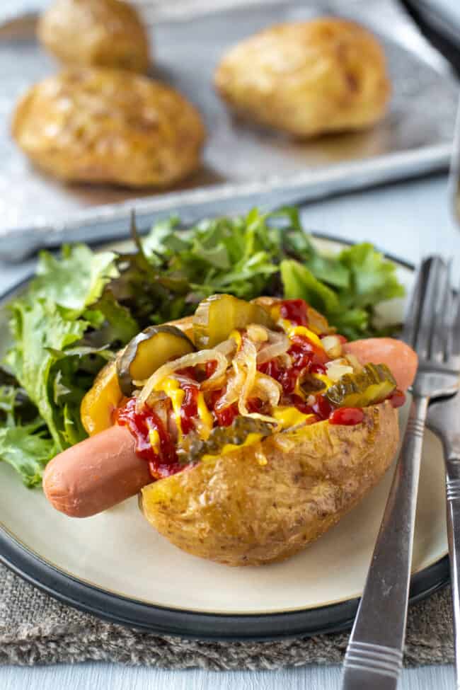 A baked potato topped with a hot dog sausage, ketchup, and fried onions.
