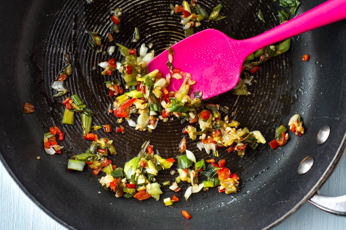 Finely chopped red chilli, spring onions and garlic being cooked in a frying pan.