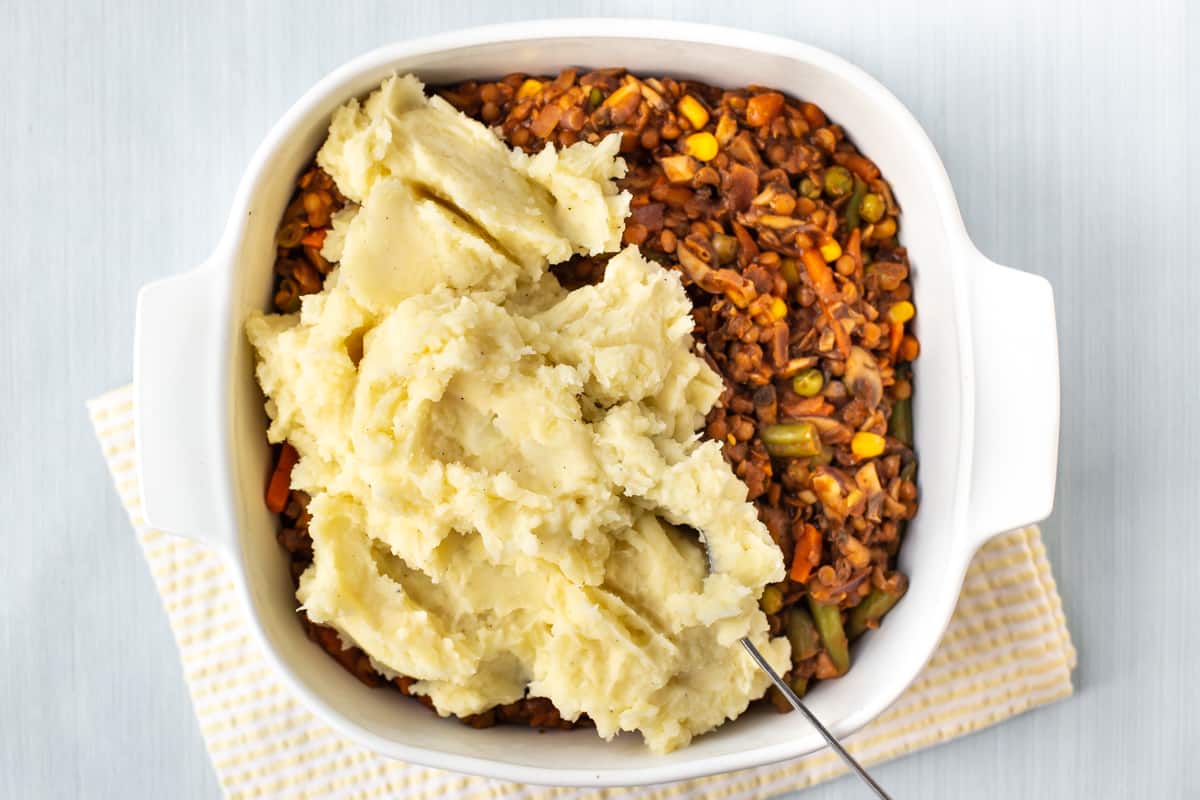 Mashed potatoes being dolloped on top of shepherd's pie filling in a dish.