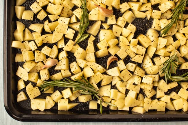 Raw potato cubes on a baking tray with fresh rosemary and garlic.