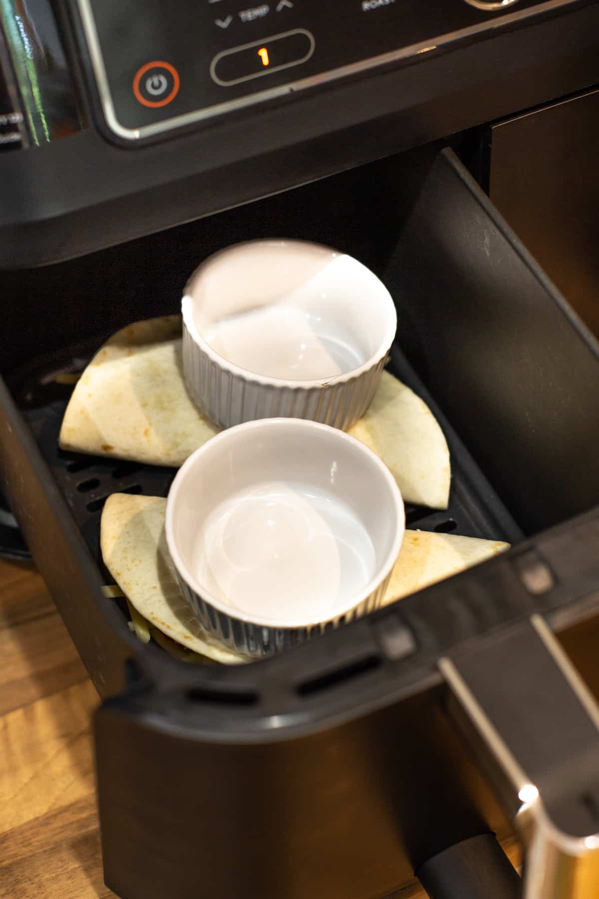Quesadillas in an air fryer being weighed down with small ramekins.