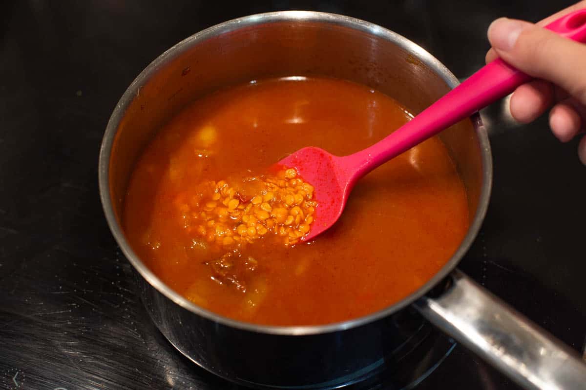 A hand stirring lentils cooking in liquid in a saucepan.