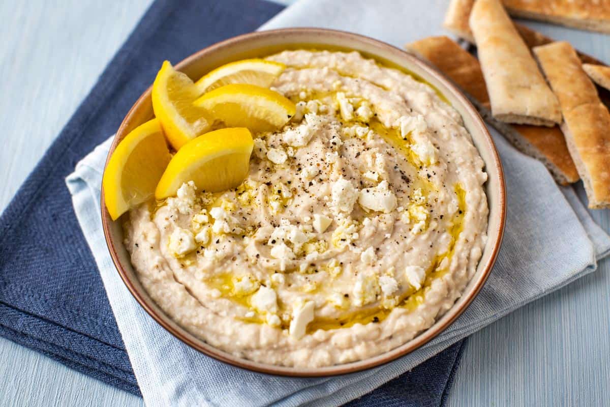 White bean hummus in a bowl topped with crumbled feta and lemon wedges.