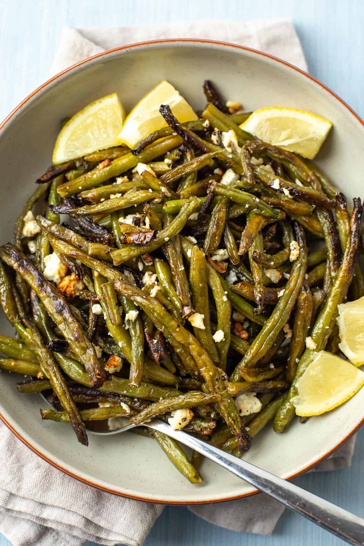 A bowl of roasted green beans with crumbled feta cheese and lemon wedges.