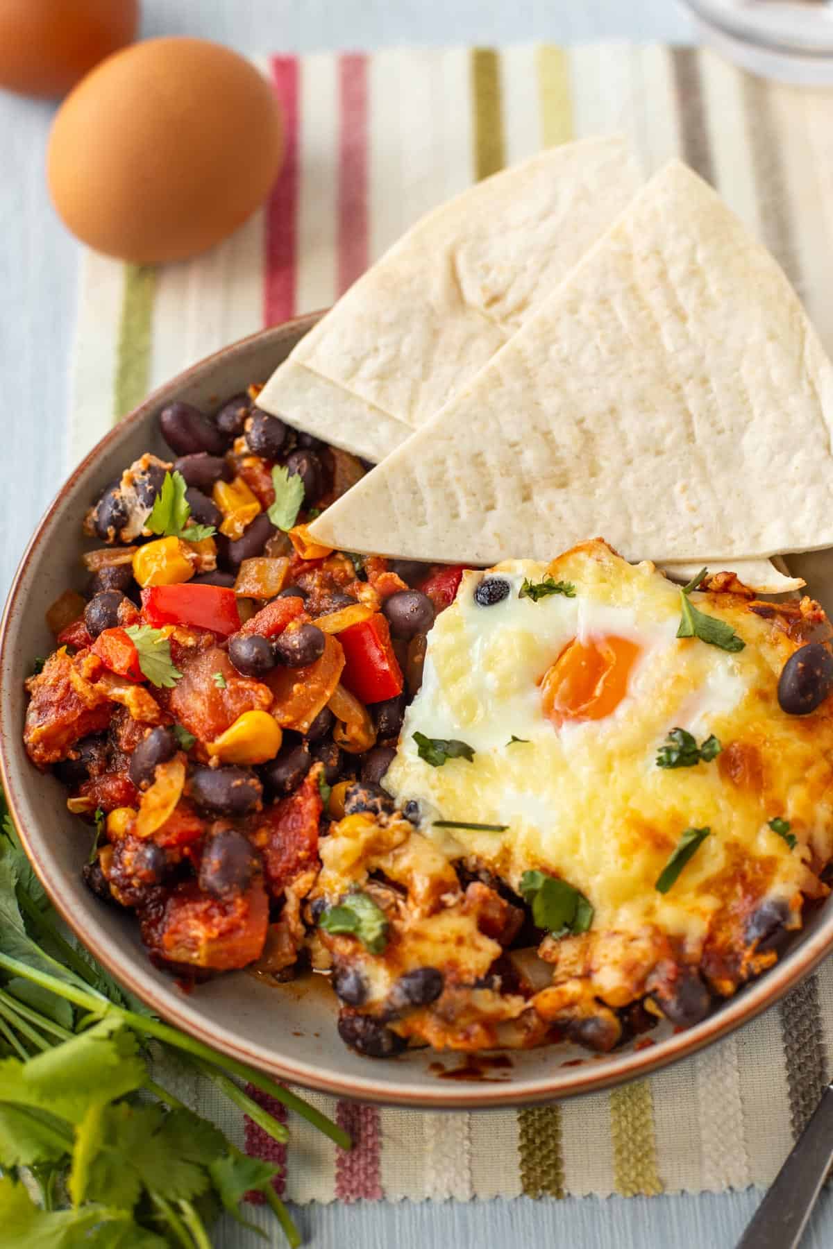 A portion of Mexican baked eggs in a bowl, with bean chilli and flour tortillas.